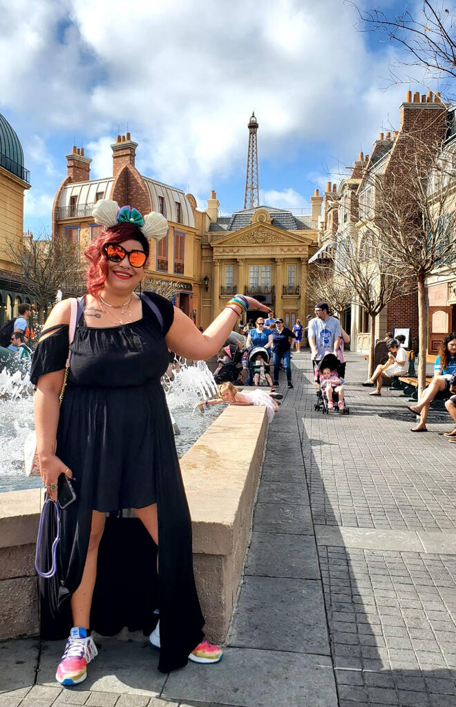 Epcot: France 🗼 a photograph of Caitlin K-S, wearing The Little Mermaid themed mouse ears, posing in front of the Eiffel Tower replica in Walt Disney World